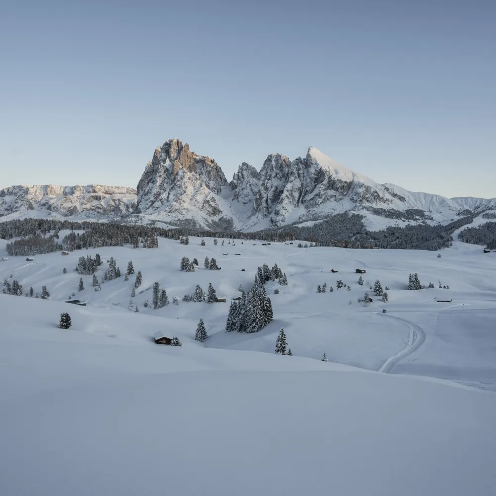 Verschneite Seiser Alm mit Almhütten und den Gipfeln der Dolomiten im ersten Morgenlicht.