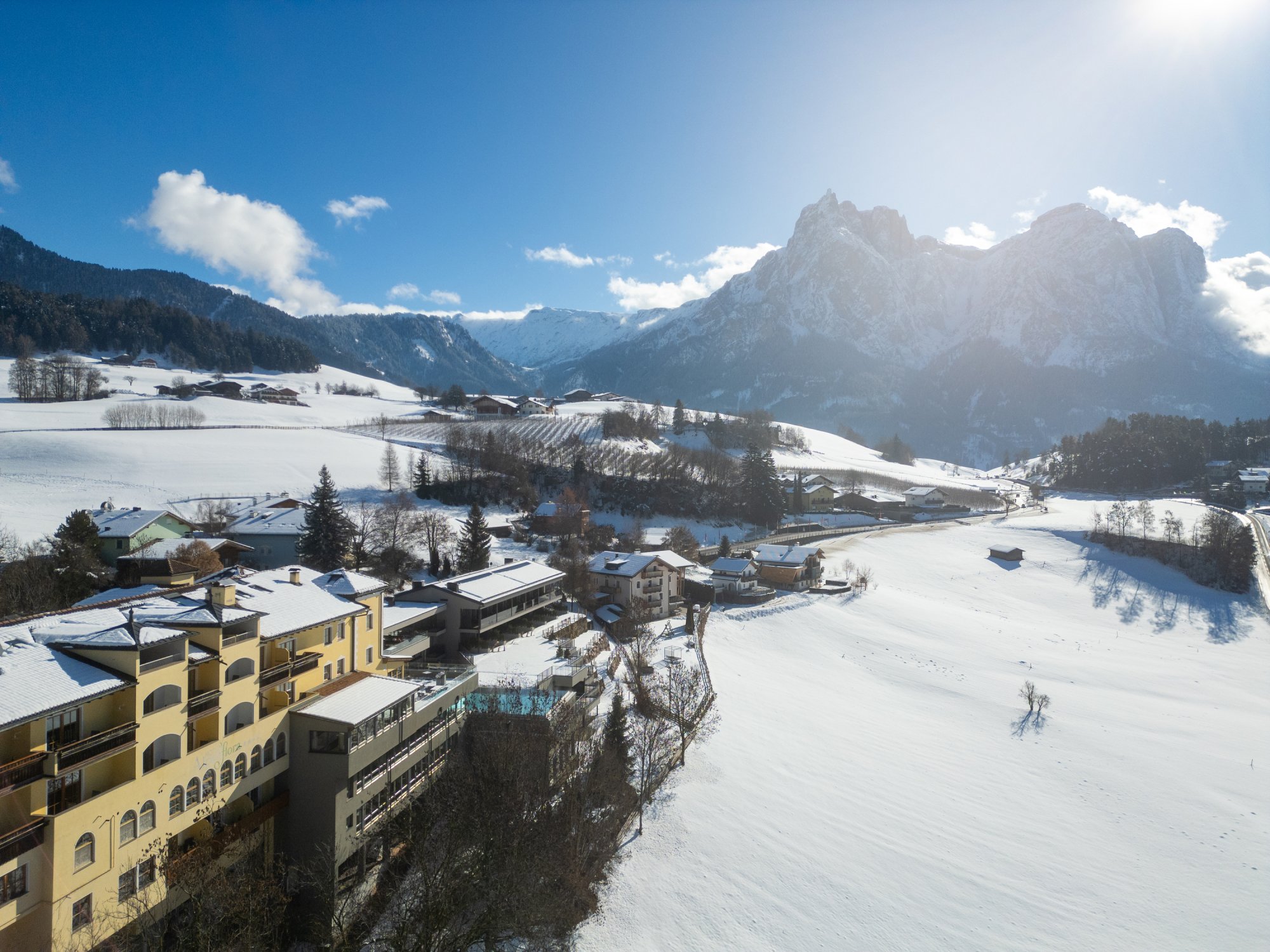 Wellnesshotel Alpenflora im Winter mit verschneiter Landschaft, Balkonen und Bergpanorama unter blauem Himmel.