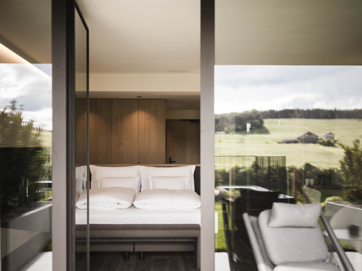 Hotelzimmer mit Doppelbett, Holzelementen und Panoramafenster zur Bergwiese bei Kastelruth