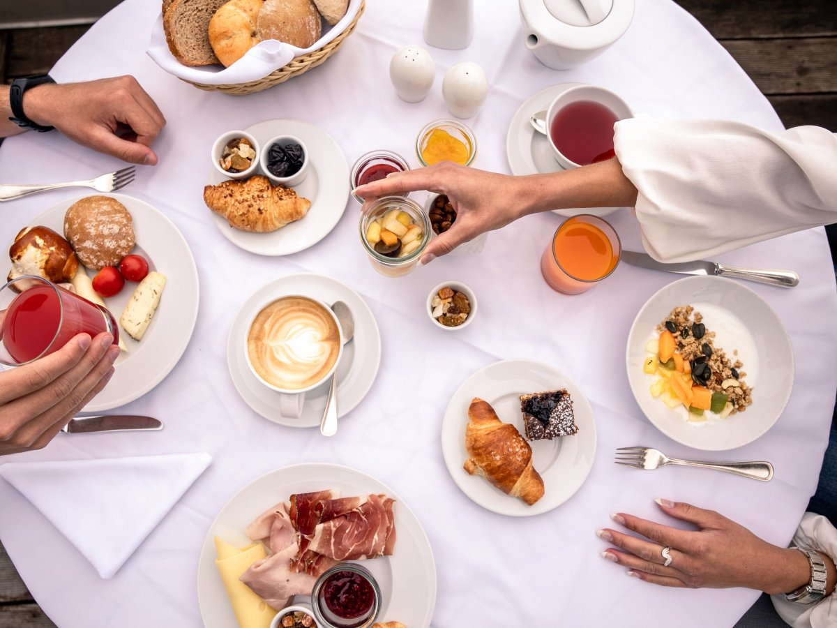Reich gedeckter Frühstückstisch von oben mit Cappuccino, Croissants, Käse und Südtiroler Speck