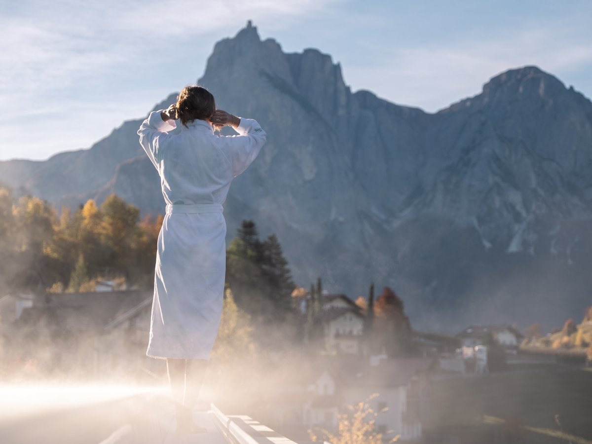 Frau im Bademantel steht im aufsteigenden Dampf am Außenpool vor herbstlich gefärbten Bäumen.