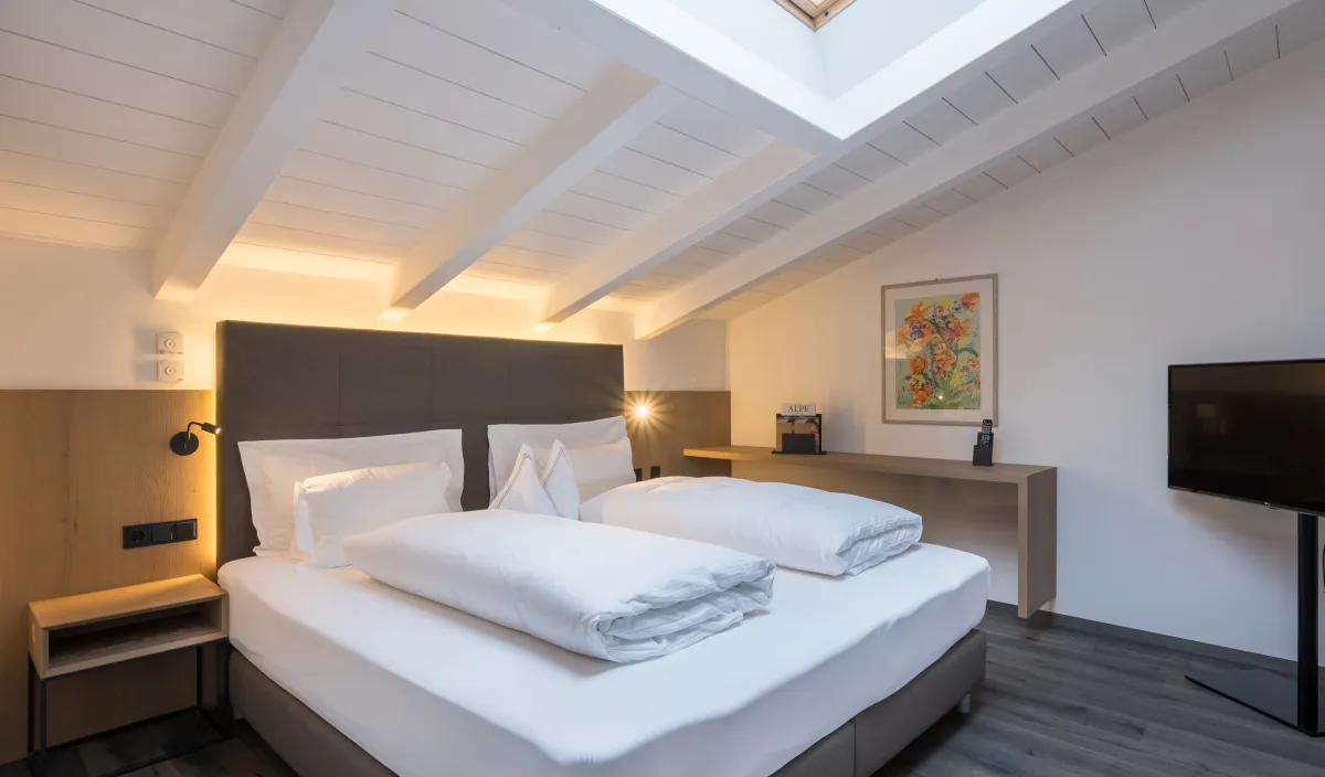 Schlafbereich der Attico Suite im Wellnesshotel Alpenflora mit Doppelbett, Dachfenster und weißer Holzbalkendecke.