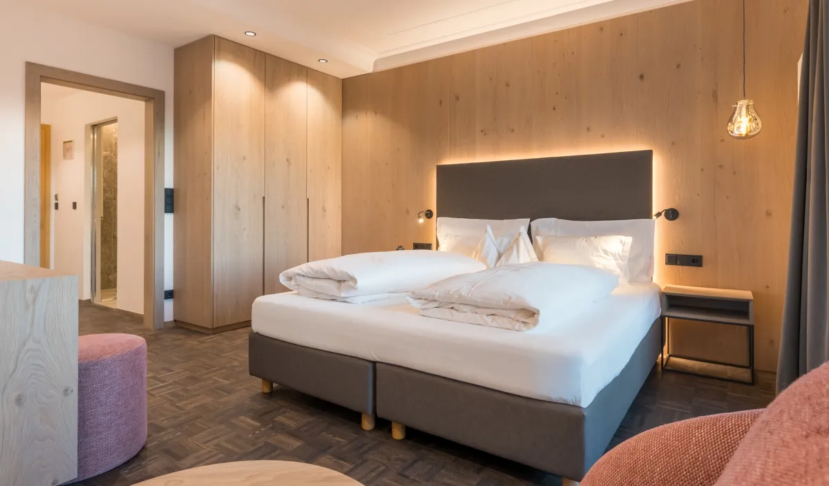 Doppelzimmer Panorama im Wellnesshotel Alpenflora mit Doppelbett, Holzschrank, Holzwandverkleidung und rosa Sitzpouf.