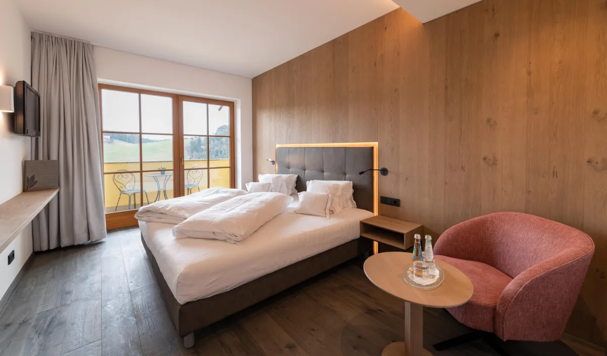 Doppelbett mit gepolstertem Kopfteil, Holzwandverkleidung, rosa Sessel und Balkontür mit Blick ins Grüne.