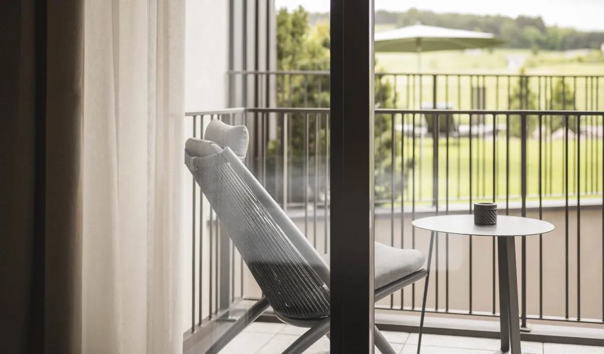 Blick vom Zimmer auf den Balkon mit Liegestuhl, Beistelltisch und grüner Landschaft.