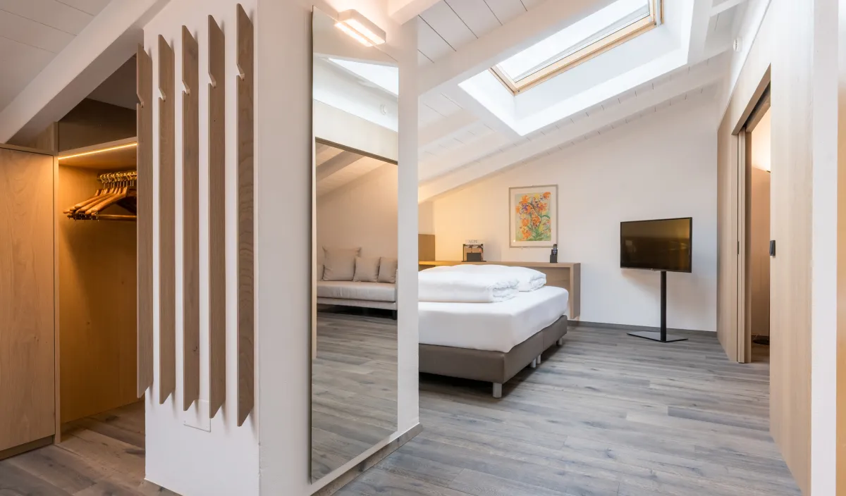 Garderobe mit beleuchtetem Kleiderschrank und Ganzspiegel, Blick auf Bett und Dachfenster der Attico Suite.