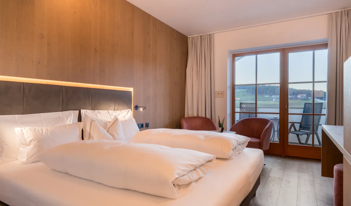 Doppelbett mit Holzwandverkleidung, Sessel in Bordeaux und Balkontür mit Blick auf Wiesen und Hügel.