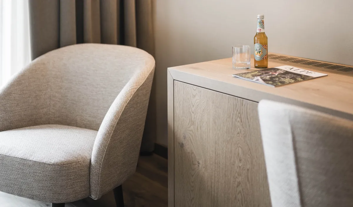 Sitzecke mit Polstersessel, Schreibtisch mit Getränk und Magazin sowie Fernseher an der Wand.