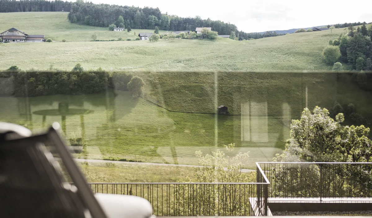 Blick vom Balkon auf grüne Wiesen und sanfte Hügel mit Liegestuhl im Vordergrund.
