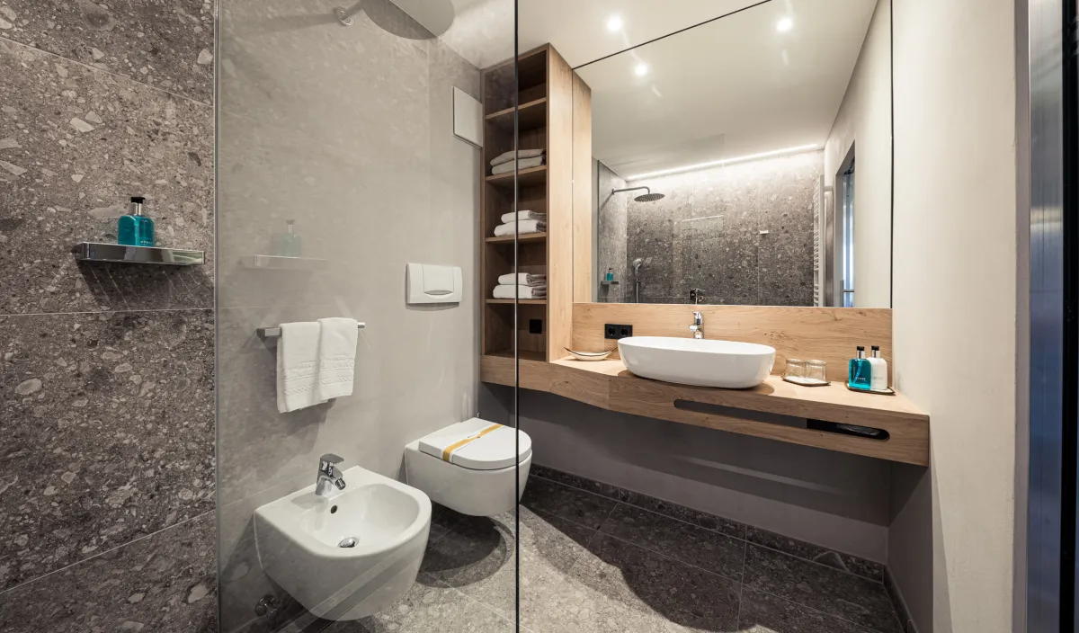 Badezimmer mit Glasdusche, Aufsatzwaschbecken auf Holzwaschtisch, Bidet, WC und Naturstein-Terrazzofliesen.