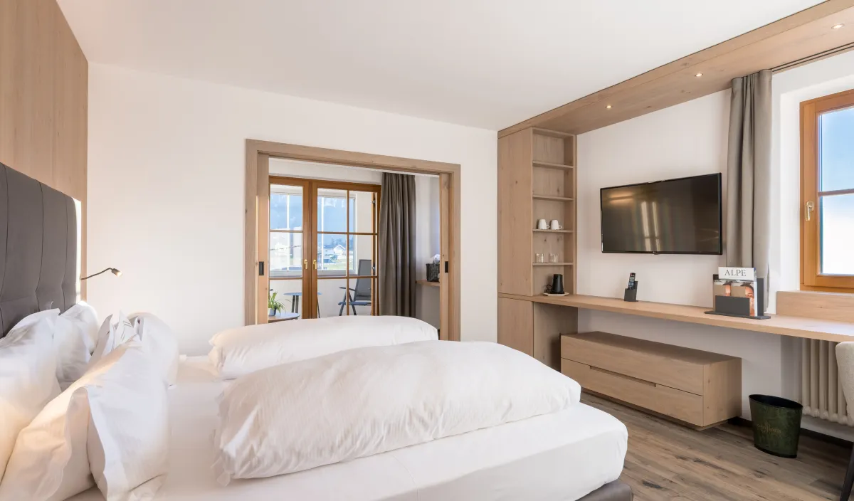 Schlafzimmer mit Doppelbett, Holzeinbauwand mit Fernseher und Regal sowie Durchgang zum Wohnraum mit Balkon.