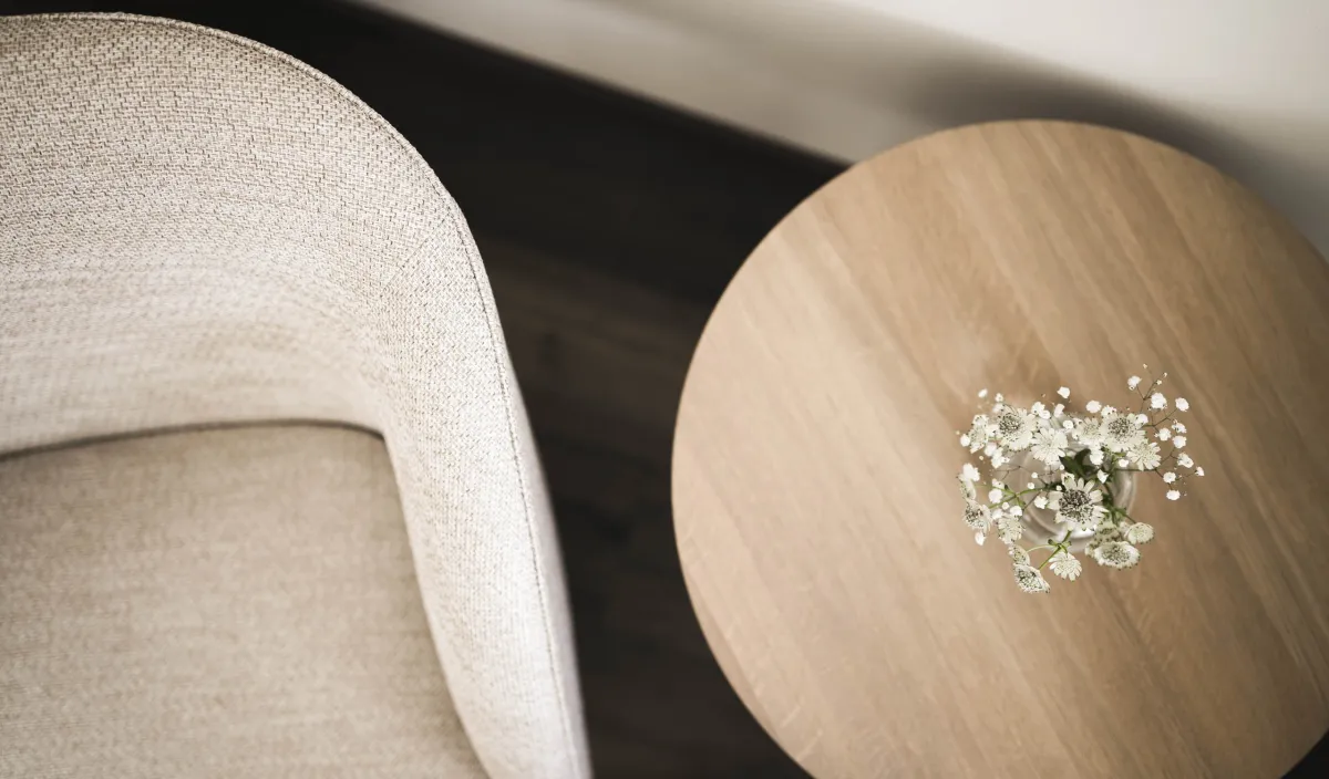 Detailaufnahme von oben mit Polstersessel, rundem Holztisch und kleinem Wiesenblumenstrauss im Glasväschen.