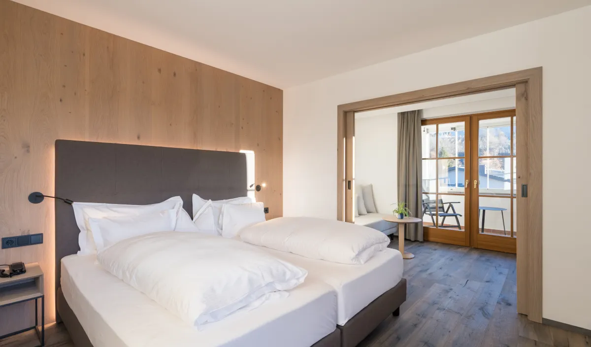 Schlafbereich mit Doppelbett, Holzwandverkleidung und Durchgang zum Wohnraum mit Sofa und Balkontür.