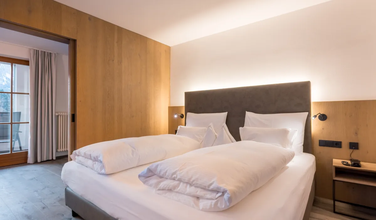 Doppelbett mit gepolstertem Kopfteil, Holzwandverkleidung und warmem Licht neben dem Durchgang zum Balkon.