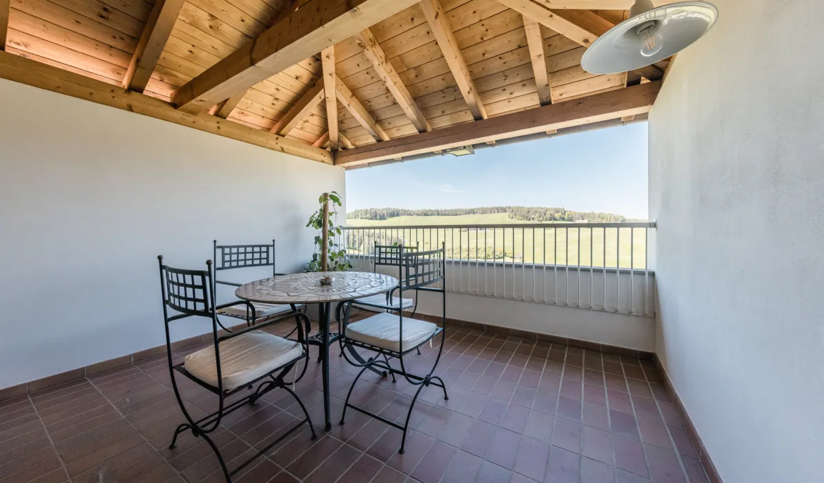 Überdachter Balkon mit Schmiedeeisen-Sitzgruppe, Holzdachstuhl und Blick auf Wiesen und Hügel.