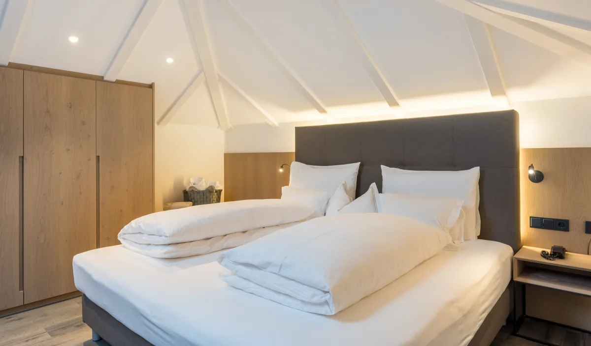 Doppelbett mit weißer Bettwäsche, gepolstertem Kopfteil und Holzschrank unter der Dachschräge.