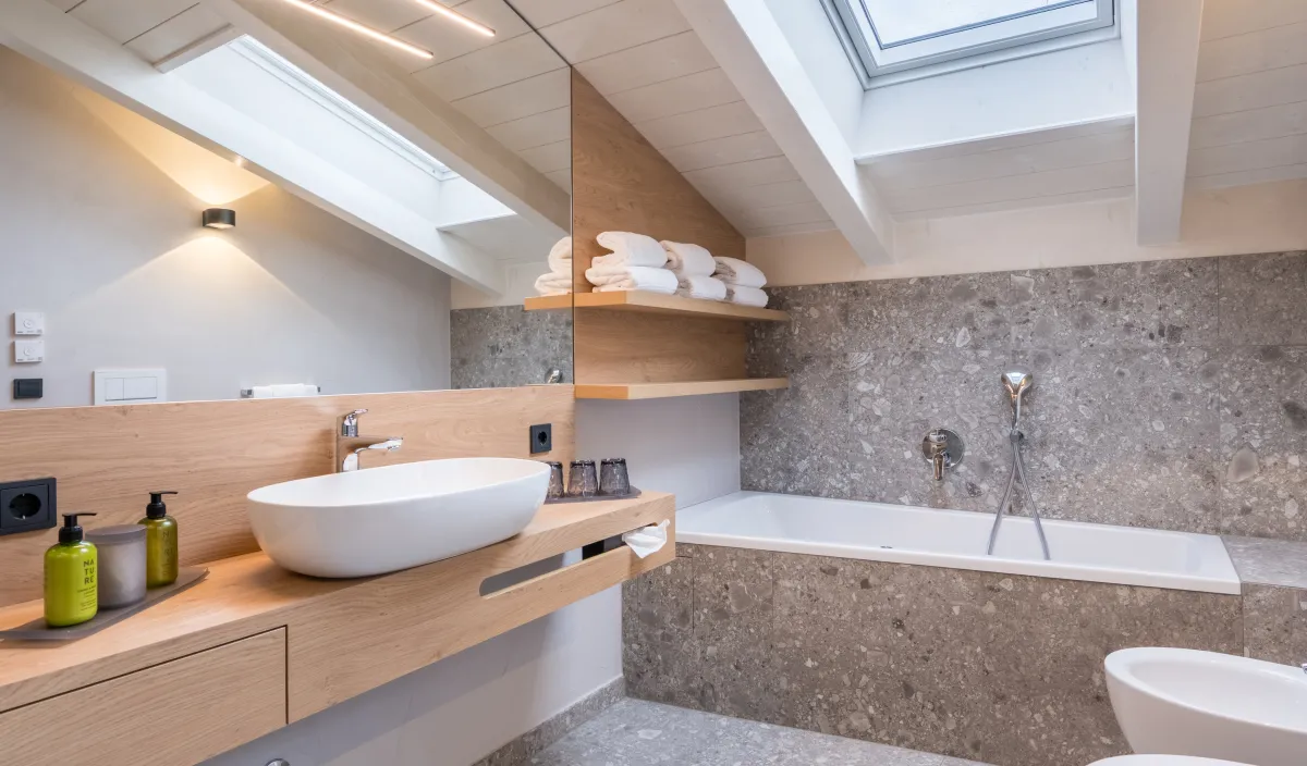 Badezimmerdetail mit Waschtisch, Badewanne, Naturstein-Terrazzofliesen und Dachfenster mit Himmelblick.
