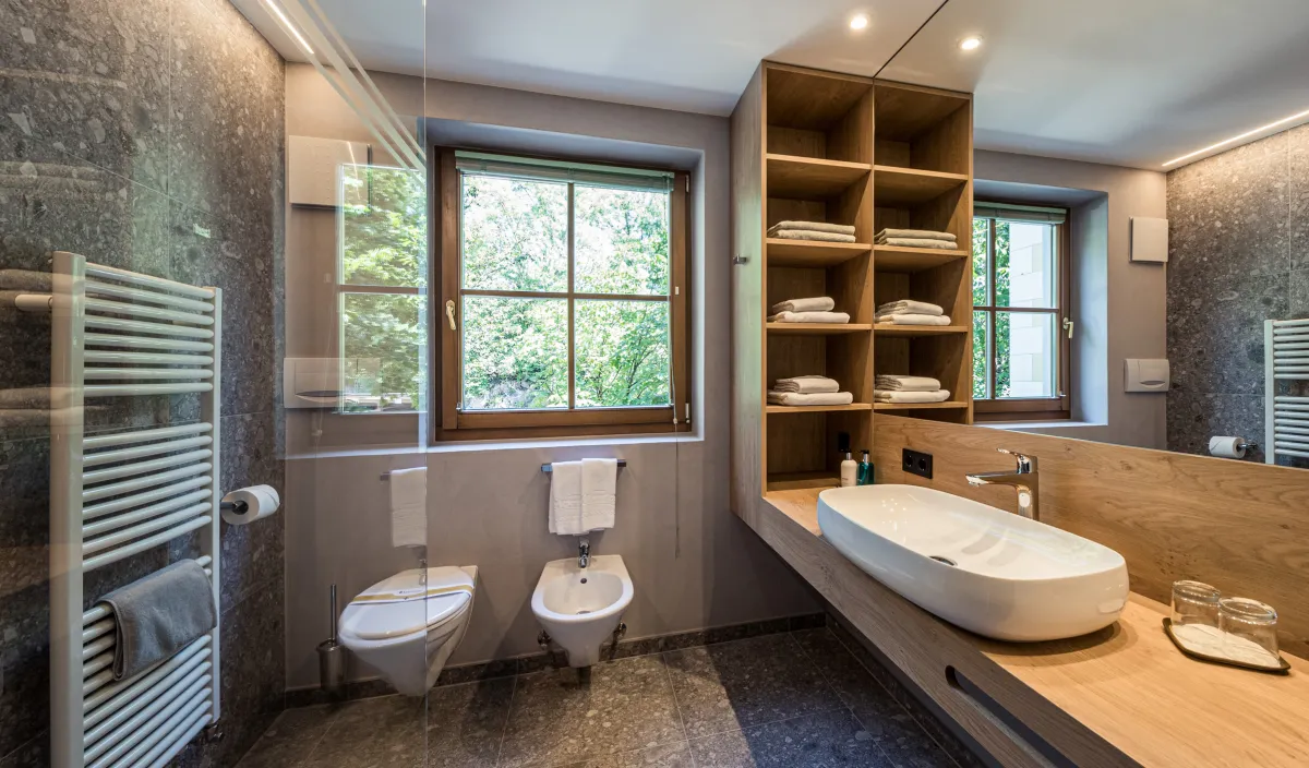 Geräumiges Badezimmer mit Waschtisch, WC, Bidet, Handtuchregalen, Fenster und Natursteinboden.