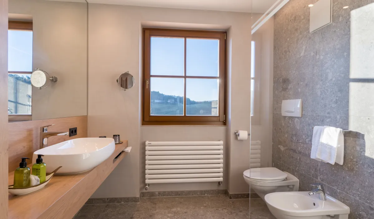 Badezimmer mit Waschtisch auf Holzplatte, grossem Spiegel, WC, Bidet und Fenster mit Blick ins Grüne.