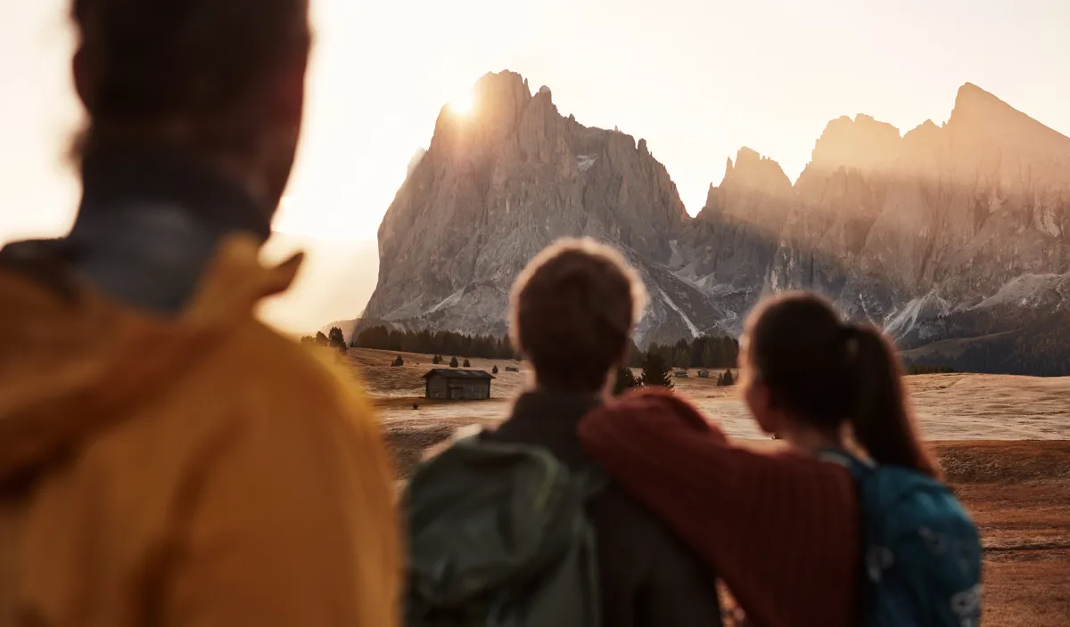 Drei Wanderer blicken auf eine Almwiese mit Holzhütte und die Gipfel der Dolomiten im Morgenlicht