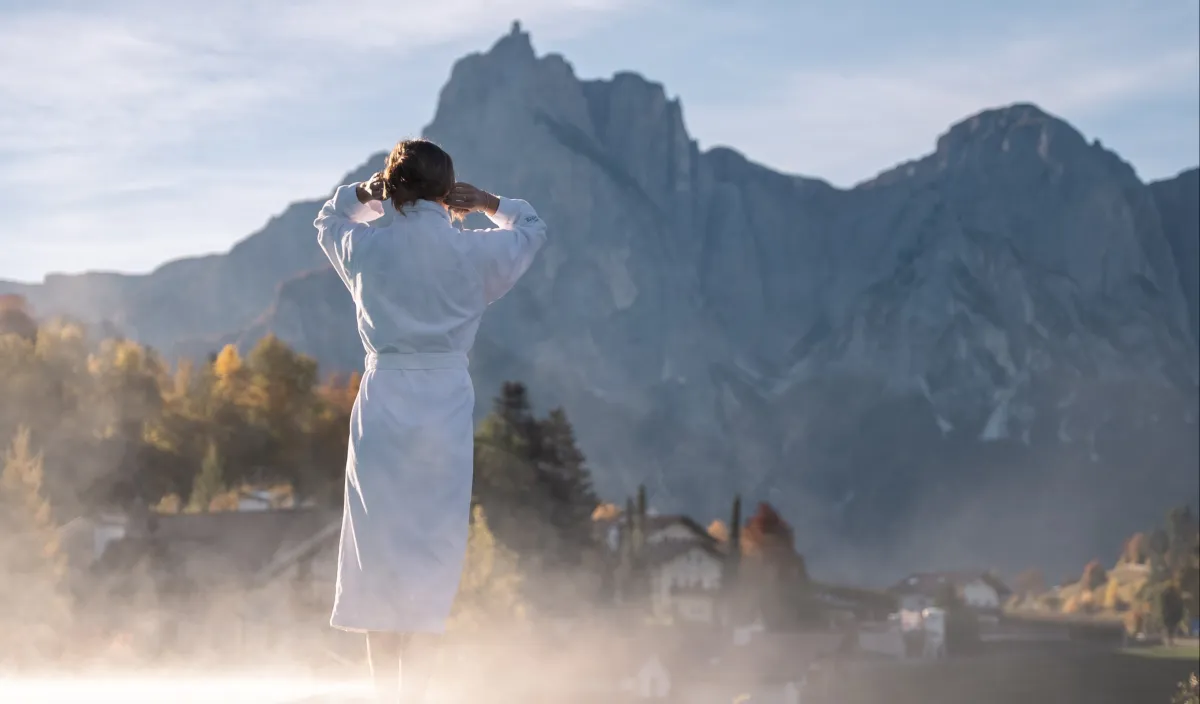 Woman in a bathrobe standing in the rising steam at the outdoor pool among autumn-coloured trees.