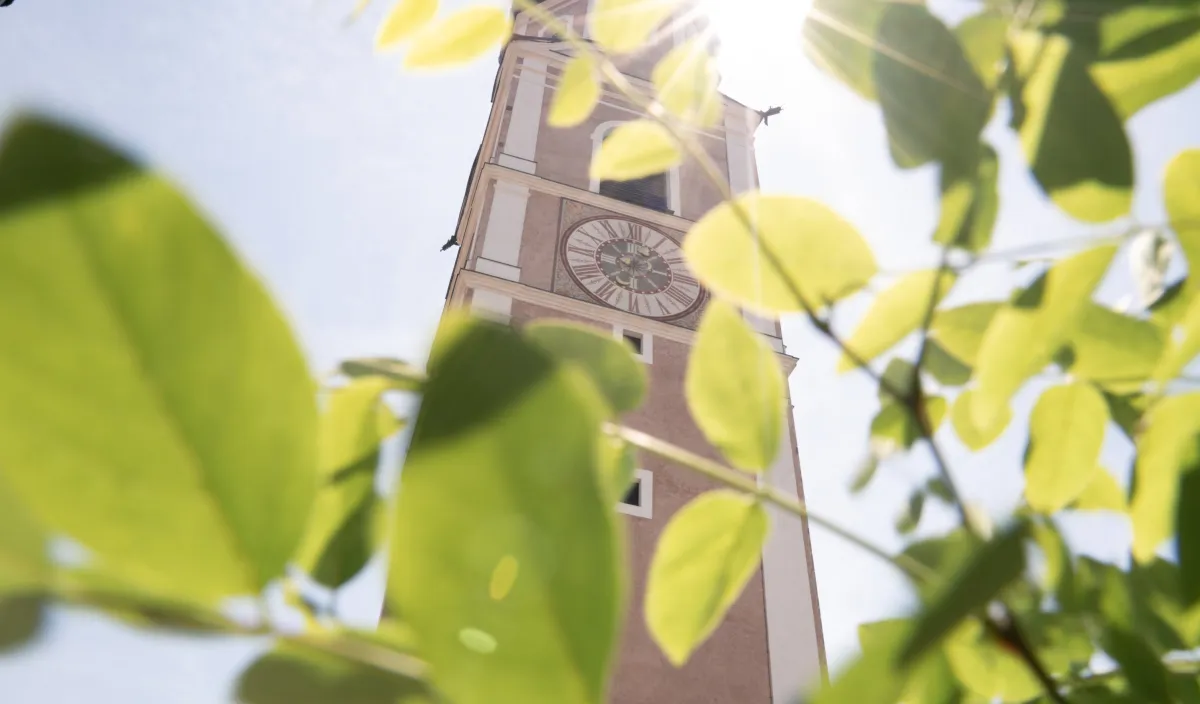 Church tower with a clock seen through green leaves in the sunlight in Castelrotto