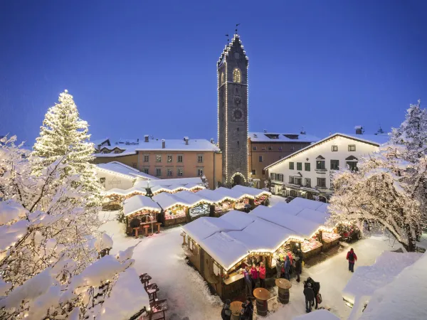 Snow-covered Christmas market with an illuminated tree and town tower at night in South Tyrol