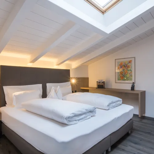 Schlafbereich der Attico Suite mit Doppelbett, weisser Holzbalkendecke, Dachfenster und Blumengemälde.