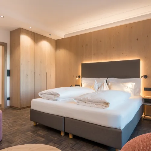 Doppelzimmer Panorama mit Doppelbett, Holzwandverkleidung, rosa Sitzpouf, Holzschrank und Pendelleuchte.